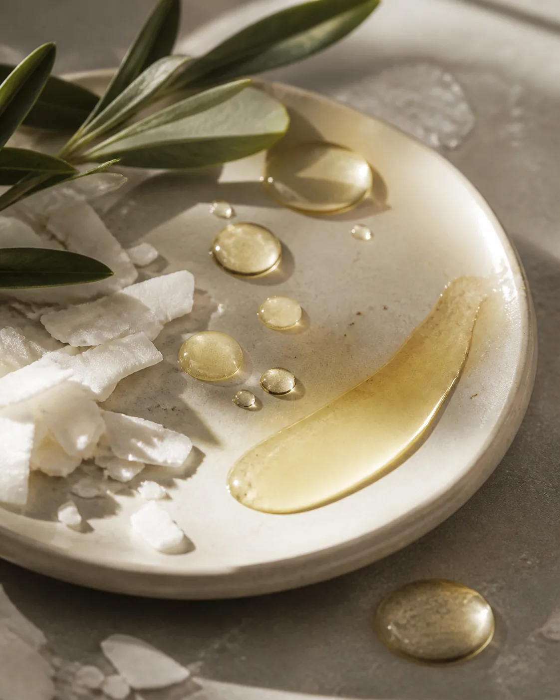 Gotas de aceite dorado con hojas botánicas y láminas blancas sobre cerámica.